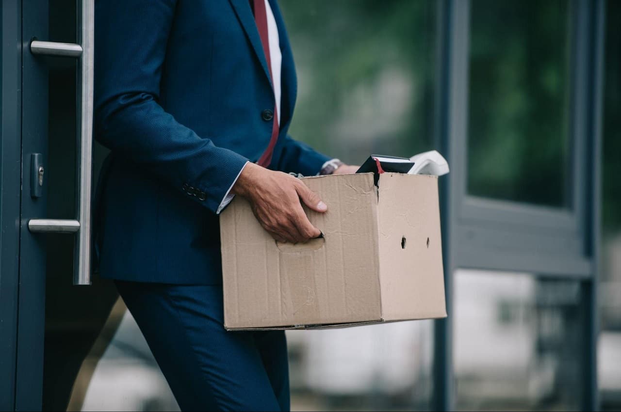 Homem carregando caixa de papelão com pertences após demissão. Ele veste terno azul-escuro e gravata vermelha (seu rosto não aparece na imagem)