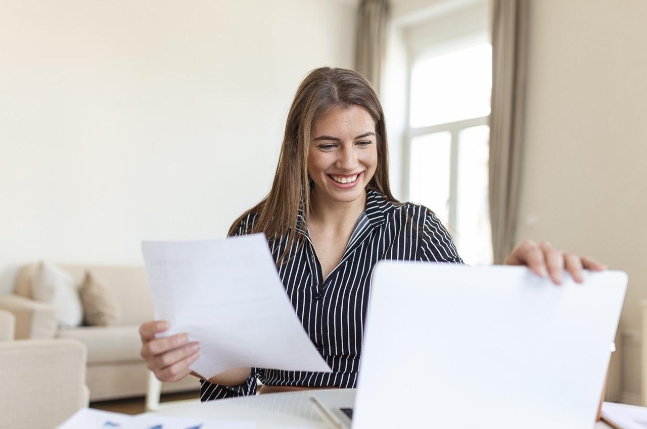 Mulher sorridente de cabelos castanhos claros, usando roupa social cinza no escritório e segurando folhas de papel.