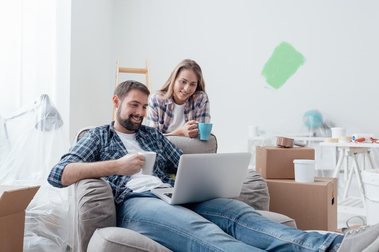 Homem sorridente sentado no sofá usa laptop em cima das pernas para acessar empréstimo para reforma. Atrás dele tem uma mulher sorridente olhando para a tela do computador. As pessoas estão na sala de sua residência em reforma.