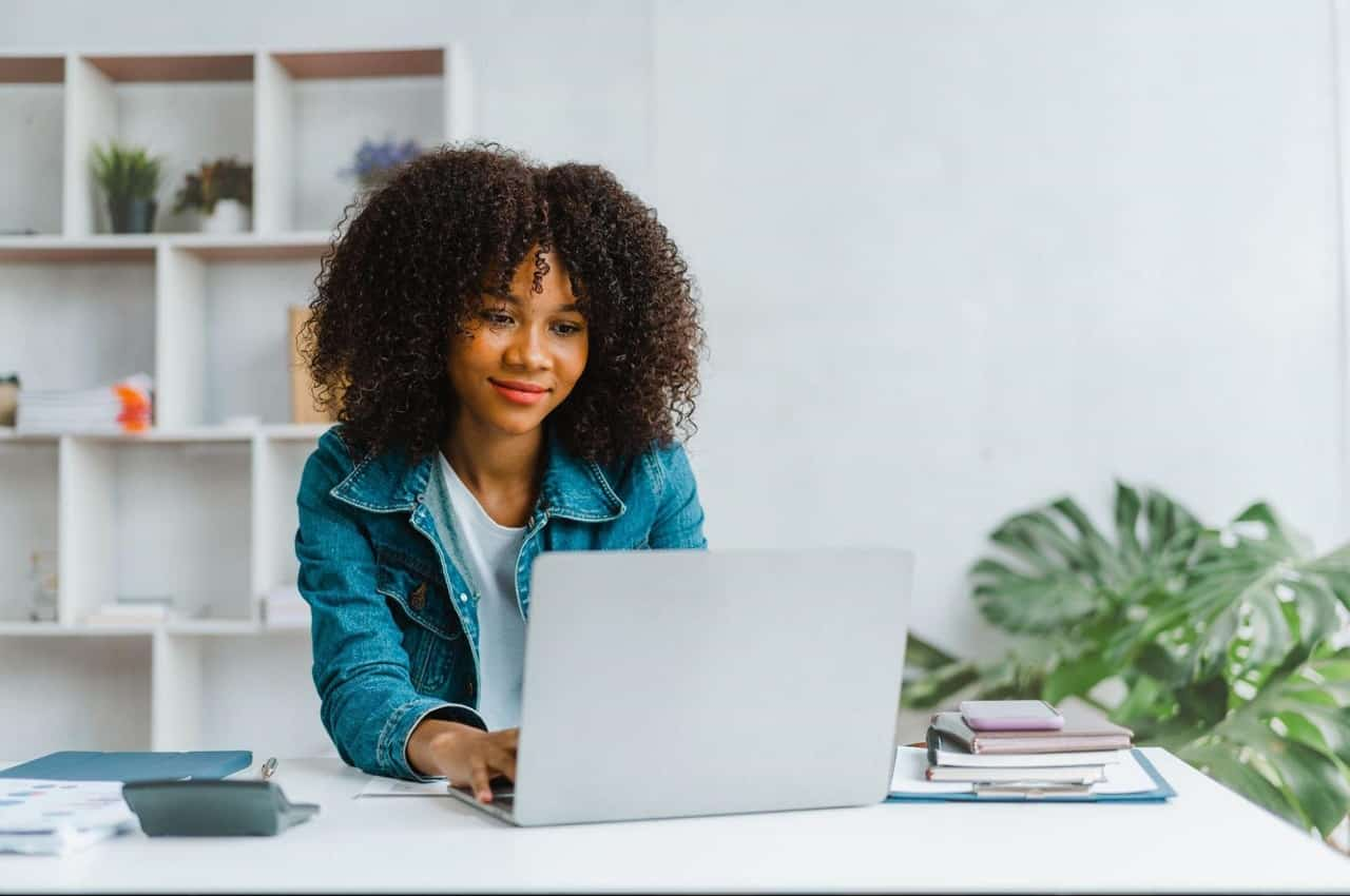 Mulher com expressão sorridente e concentrada, pesquisando análise de empréstimo pessoal online no computador. Ela está em seu escritório e utilizando o aparelho na sua mesa.