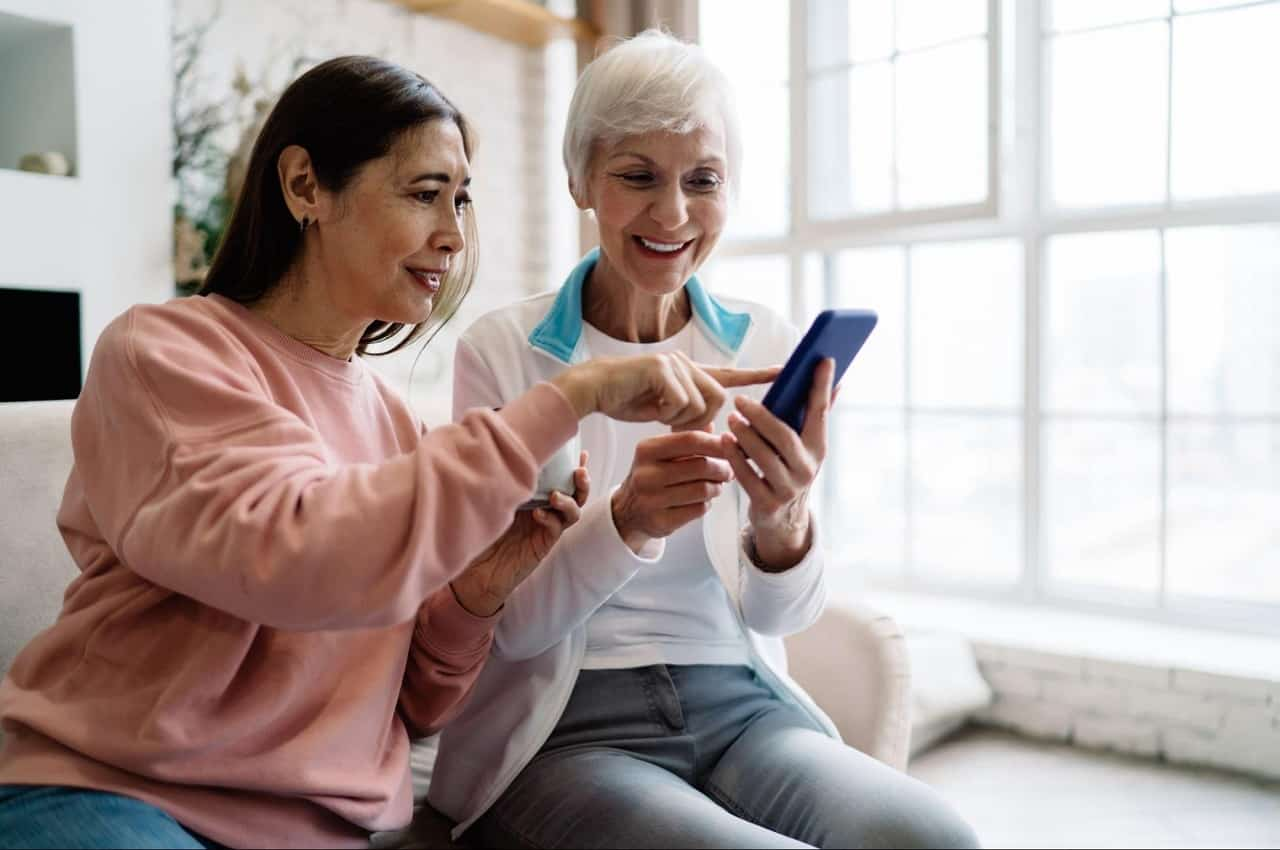 Casal sorridente de mulheres sêniores, acessando empréstimo pessoal online pelo celular. Uma das mulheres aponta para a tela do aparelho.