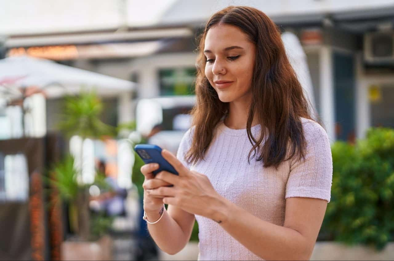 Mulher com expressão risonha, cabelos castanhos longos soltos e segurando o celular com duas mãos.