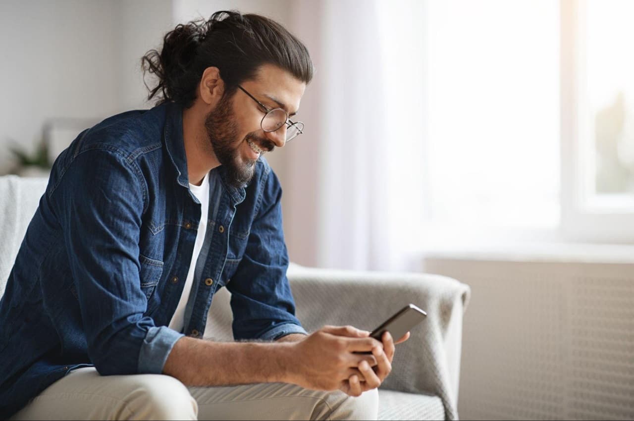 Homem sorridente com óculos de grau e utilizando celular. Ele está sentado no sofá e veste calça bege e jaqueta jeans.