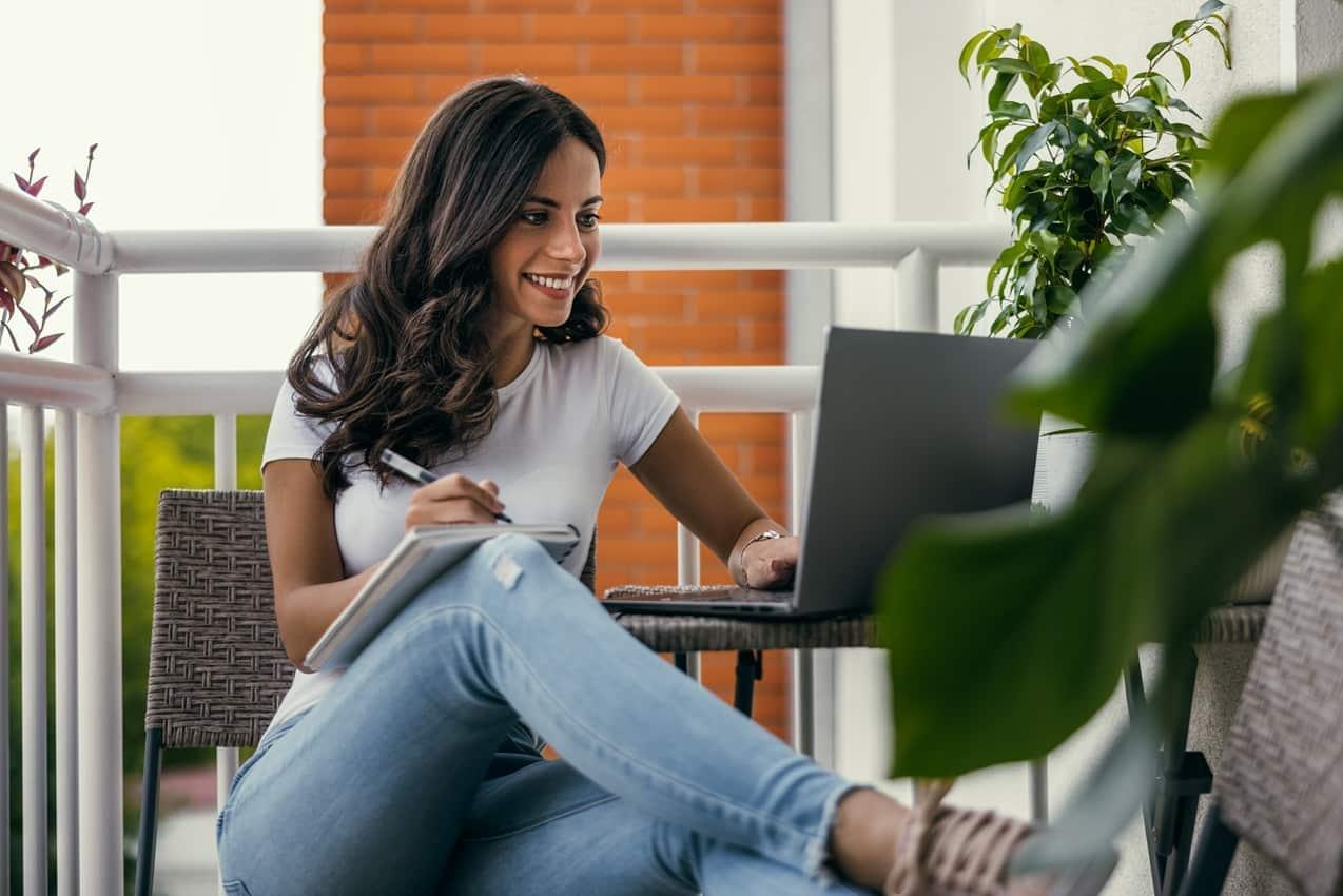 Mulher sorridente de longos cabelos lisos castanhos, está sentada em mesa de bambu e usa um laptop e um caderno para pesquisar sobre empréstimo pré-aprovado. Ela veste calça jeans e camisa básica branca.