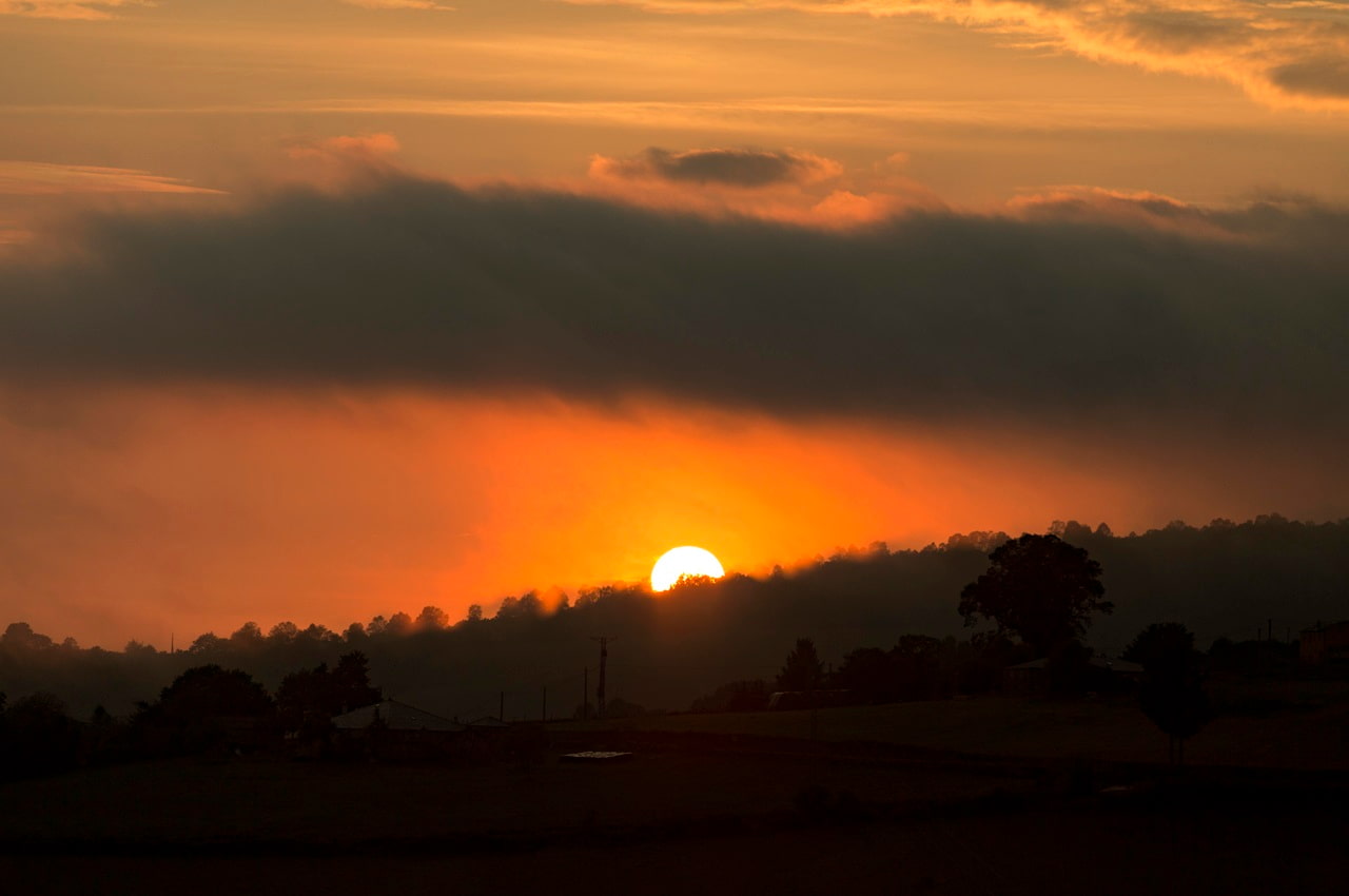 Foco em pôr do sol atrás de uma montanha cheia de árvores. O sol faz sombra na floresta e casas próximas e deixa o céu alaranjado.