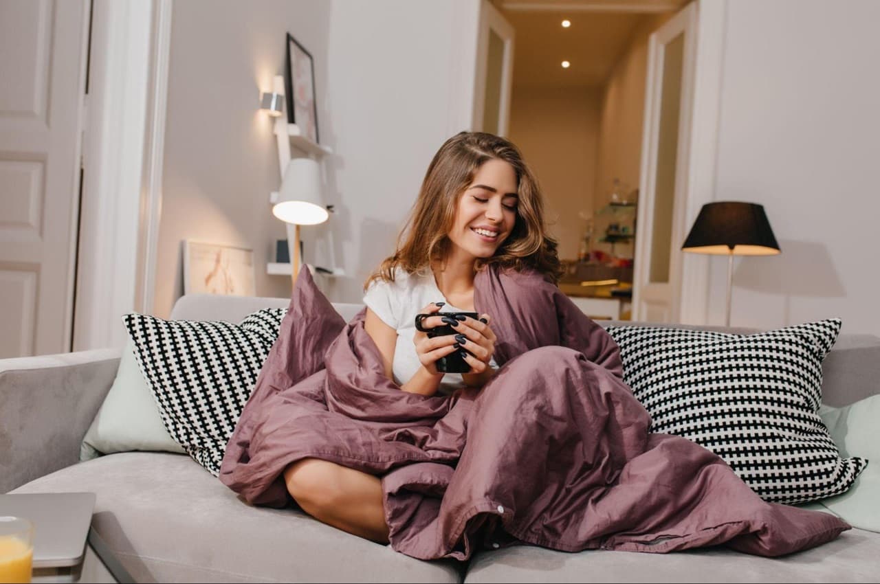 Mulher sorridente sentada no sofá de casa, segurando uma xícara de café e enrolada em um edredom rosa escuro com abajures acesos por energia solar no inverno.