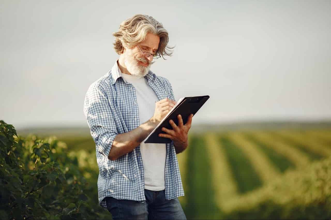 Homem de cabelos e barba grisalha com camisa xadrez azul e um tablet dentro de uma plantação rural.