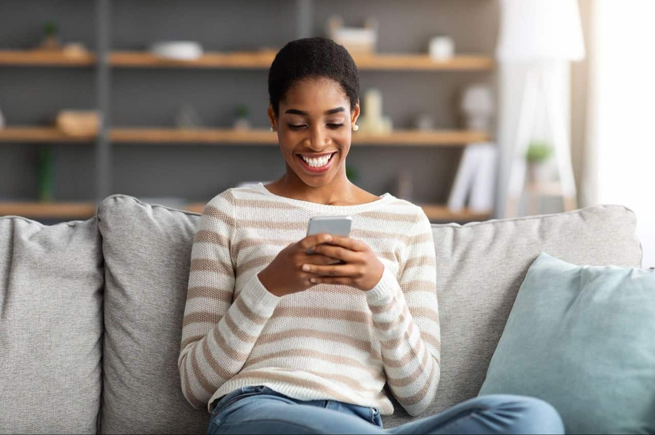 Mulher sorridente sentada no sofá de casa, digitando no celular. Ela tem cabelos curtos pretos, veste blusa listrada marrom e calça jeans.