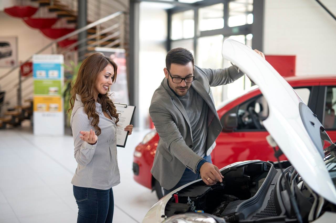 Vendedora de carros segurando planilha e mostrando estado de conservação do motor do carro usado vermelho para um cliente.