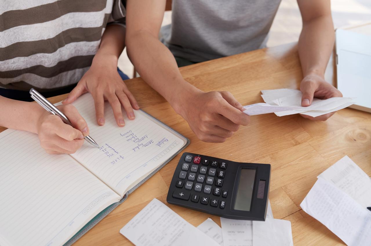 A imagem mostra duas pessoas fazendo um planejamento financeiro do mês. Elas estão escrevendo em um caderno e fazendo contas na calculadora.