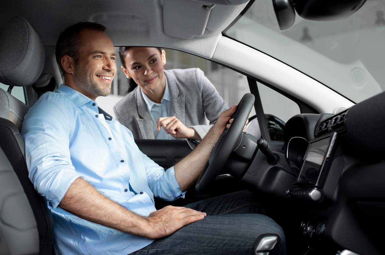 Homem sorridente fazendo test drive de veículo e vendedora da concessionária acompanhando da janela de motorista.