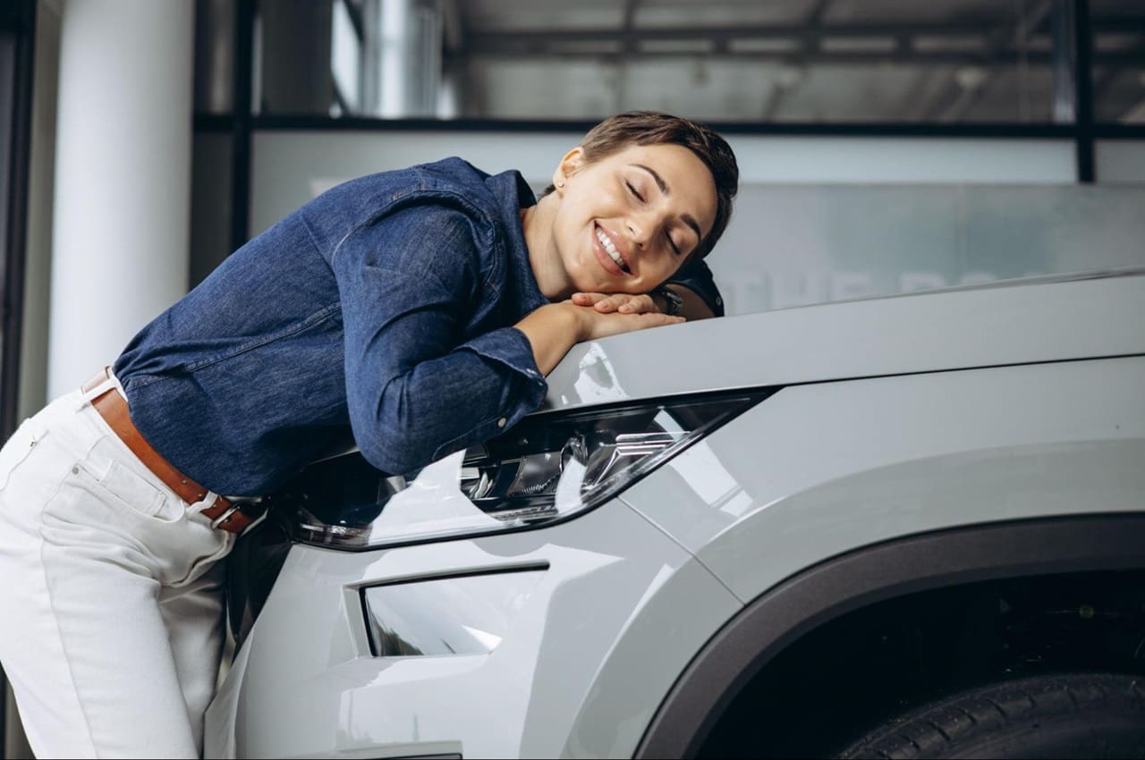 Mulher sorridente de cabelos curtos castanhos veste jaqueta azul e calça bege jeans e abraça capo de veículo financiado.