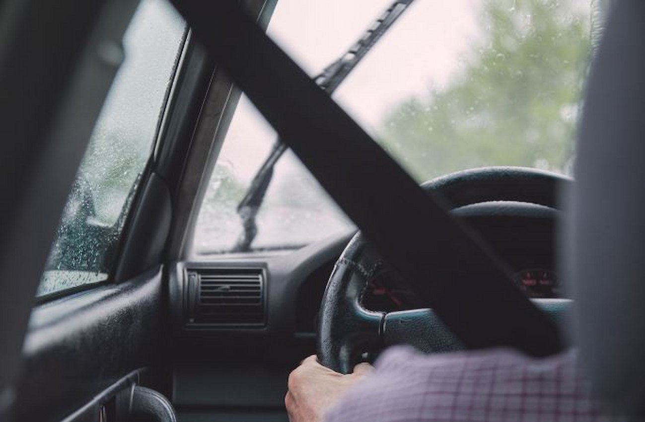 A imagem mostra a visão do banco de trás de uma pessoa dirigindo um carro.