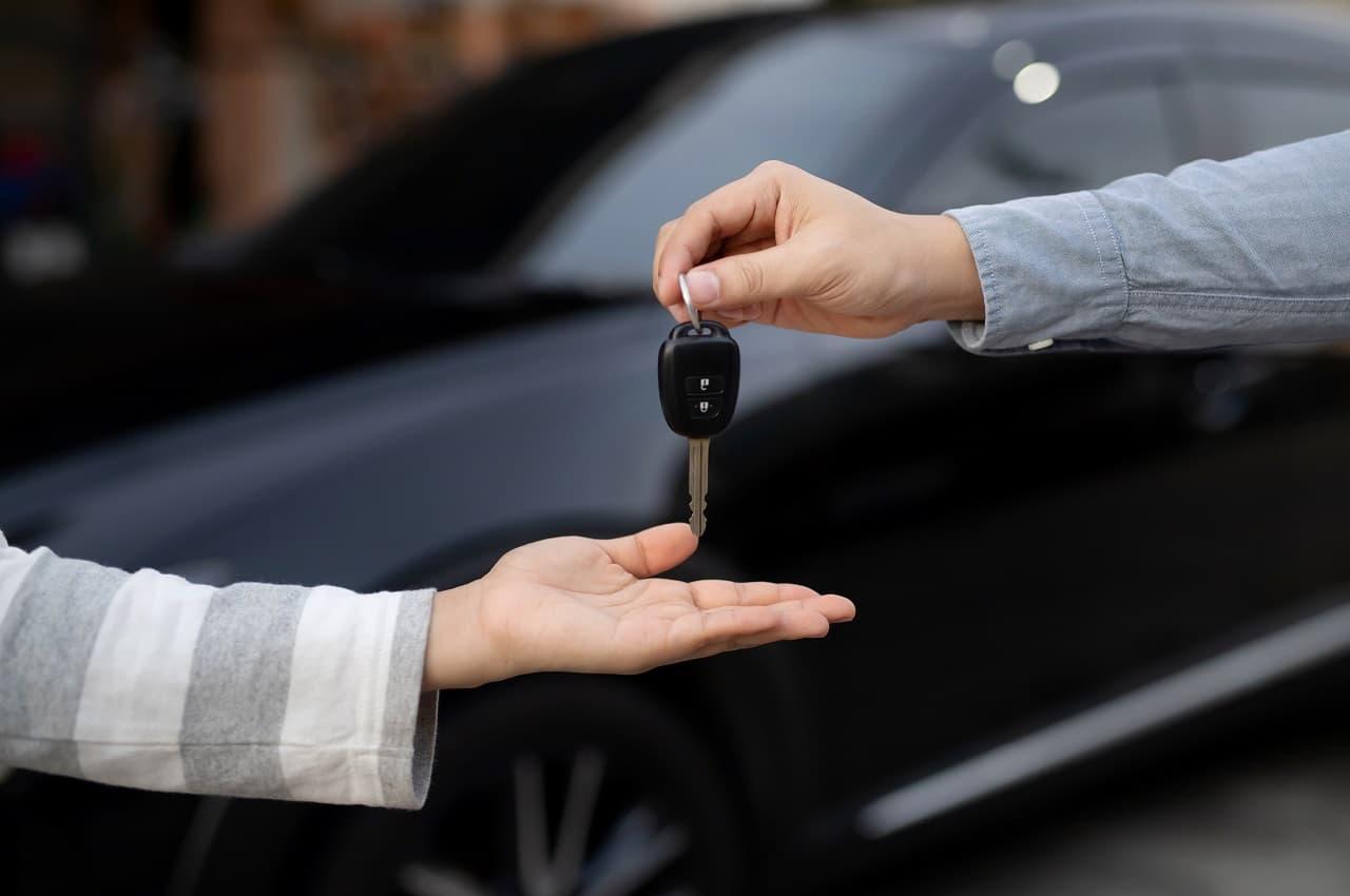 Uma mão entregando uma chave de carro para outra pessoa. Ao fundo tem um carro preto borrado pela foto.