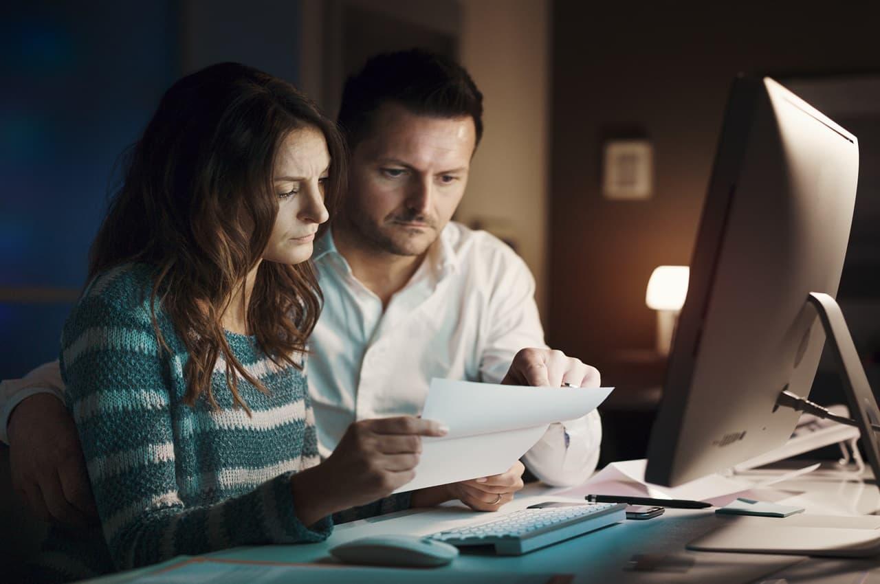 Casal de mulher e homem segura folha de papel nas mãos com as expressões preocupadas. O homem aponta para uma informação no papel e a mulher olha para a tela de um computador de mesa.