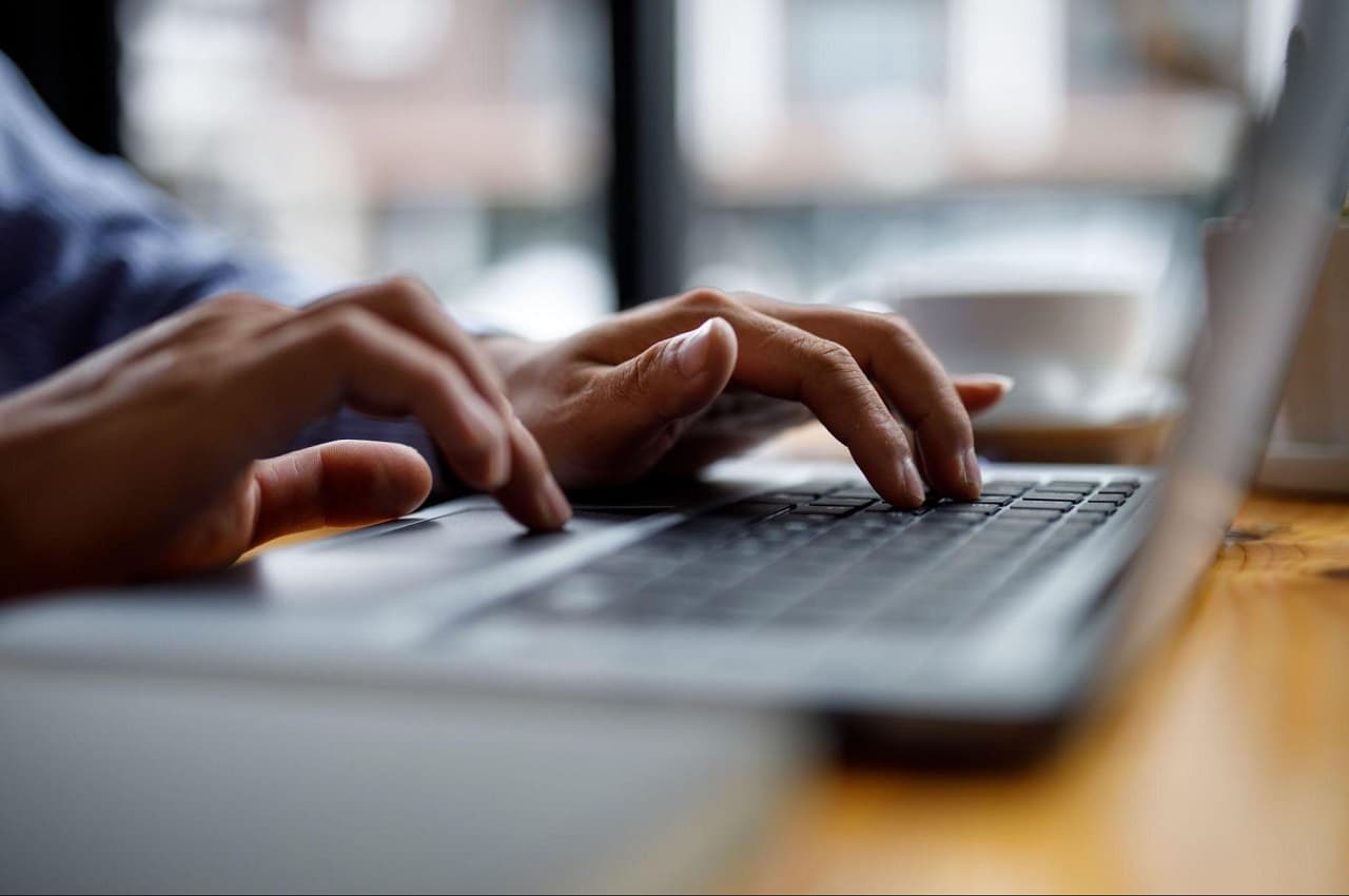 Foco em mãos digitando em teclado de laptop que está sobre uma mesa de madeira. A mesa fica ao lado de uma janela grande.