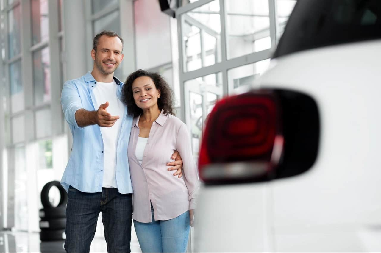 A imagem mostra um casal sorridente, olhando para um carro dentro de uma concessionária.