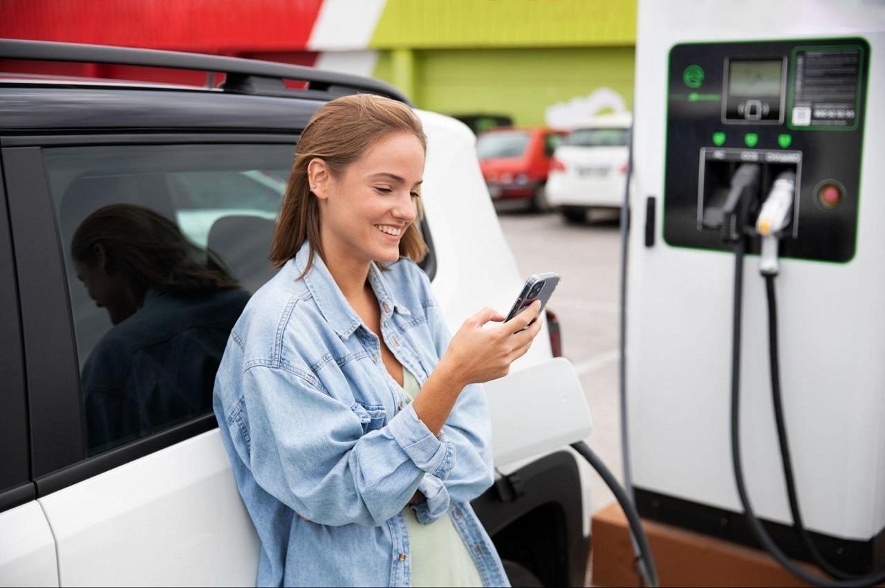 Mulher sorridente digitando no celular enquanto seu carro elétrico carrega no totem do posto de gasolina.