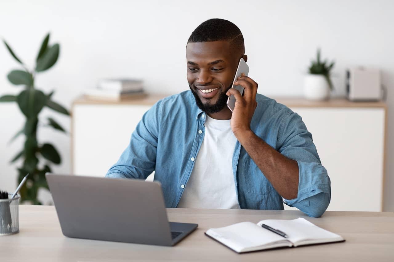 Homem negro sorridente de cabelos curtos, falando ao celular e digitando, ao mesmo tempo, no computador. Ele veste camisa branca básica, jaqueta jeans e está no escritório do trabalho.