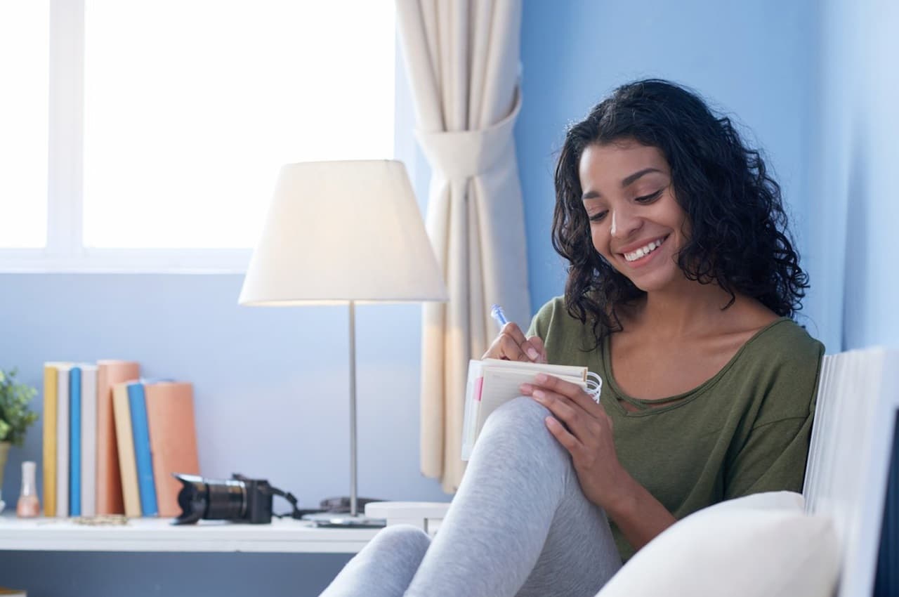 Mulher sorridente, sentada no sofá e escrevendo em um bloco de notas os gastos mensais.