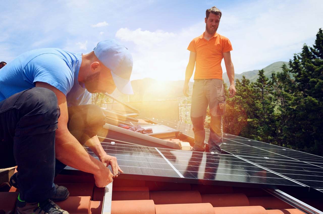 Homem agachado e com roupa de proteção instalando placa solar no telhado de uma casa, enquanto outro homem está em pé.