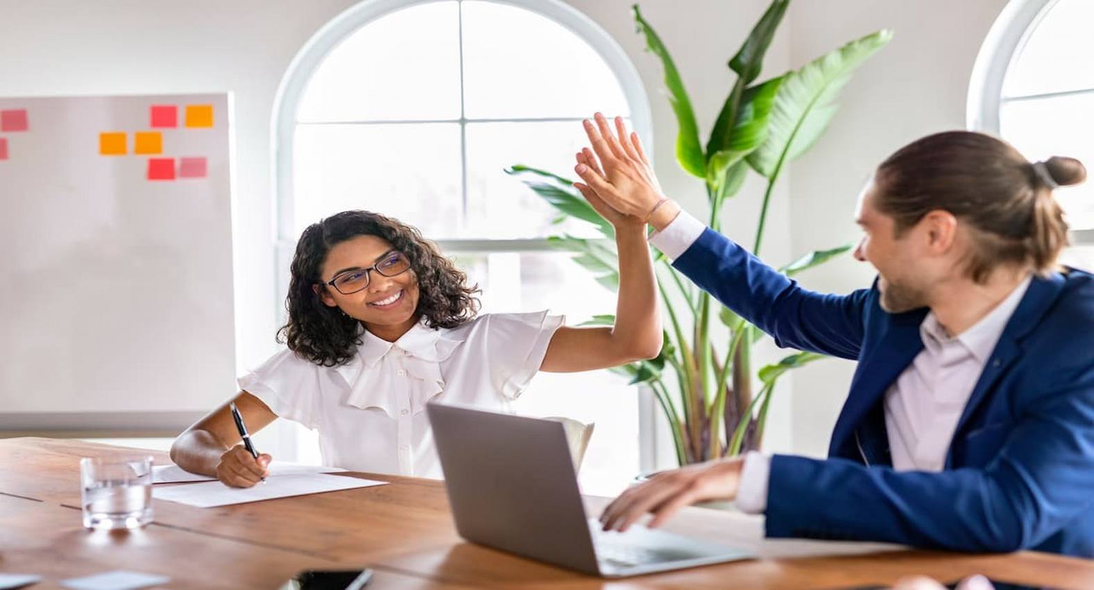 A imagem mostra duas pessoas sentadas à uma mesa, batendo suas mãos. 