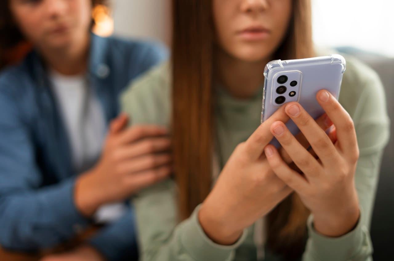 Duas pessoas sentadas lado a lado no sofá. Uma das mulheres com blusa verde segura celular com expressão preocupada, enquanto a outra mulher de blusa azul conforta ela.