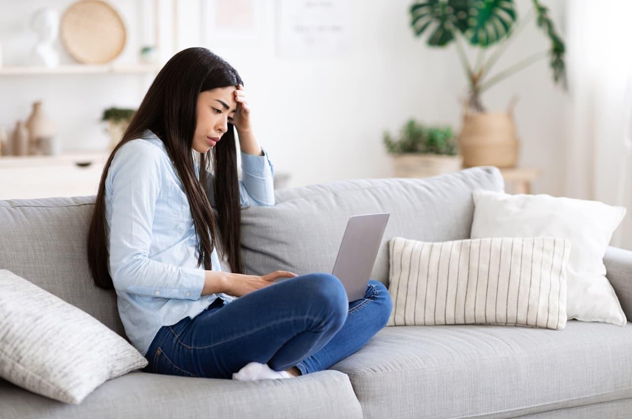 Mulher com expressão preocupada, apoia a mão na testa e pesquisa por golpe do empréstimo consignado no computador sob seu colo. Ela veste calça jeans, camisa básica branca e meias.