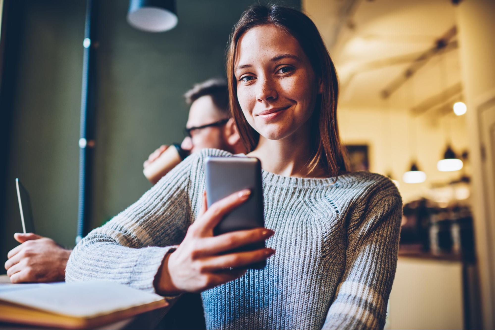 A imagem mostra uma mulher sorridente, em um estabelecimento. Ela está segurando o seu celular.