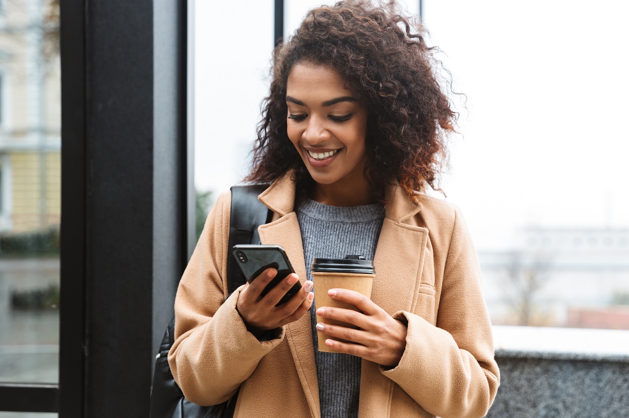 A imagem mostra uma mulher sorridente, usando seu celular e segurando um copo de café.