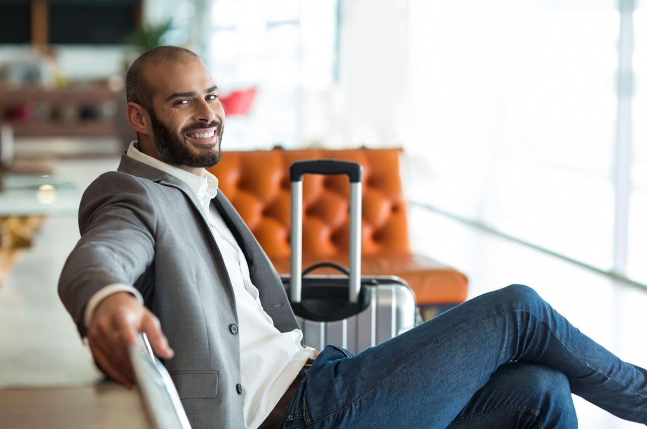 Um homem sorrindo enquanto se reclina em uma cadeira, usando um terno cinza e uma camisa branca. Ao lado dele, há uma mala de viagem. O fundo apresenta um ambiente moderno e bem iluminado, com um sofá de couro laranja ao fundo.