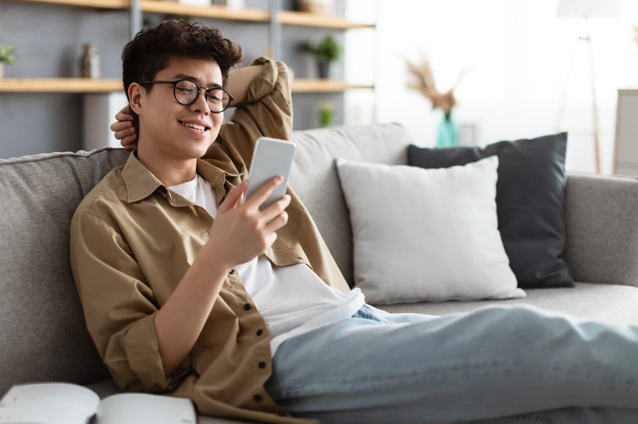 Homem usando camiseta branca básica e camisa social marrom por cima. Ele está sentado em um sofá, sorridente e usando seu celular.