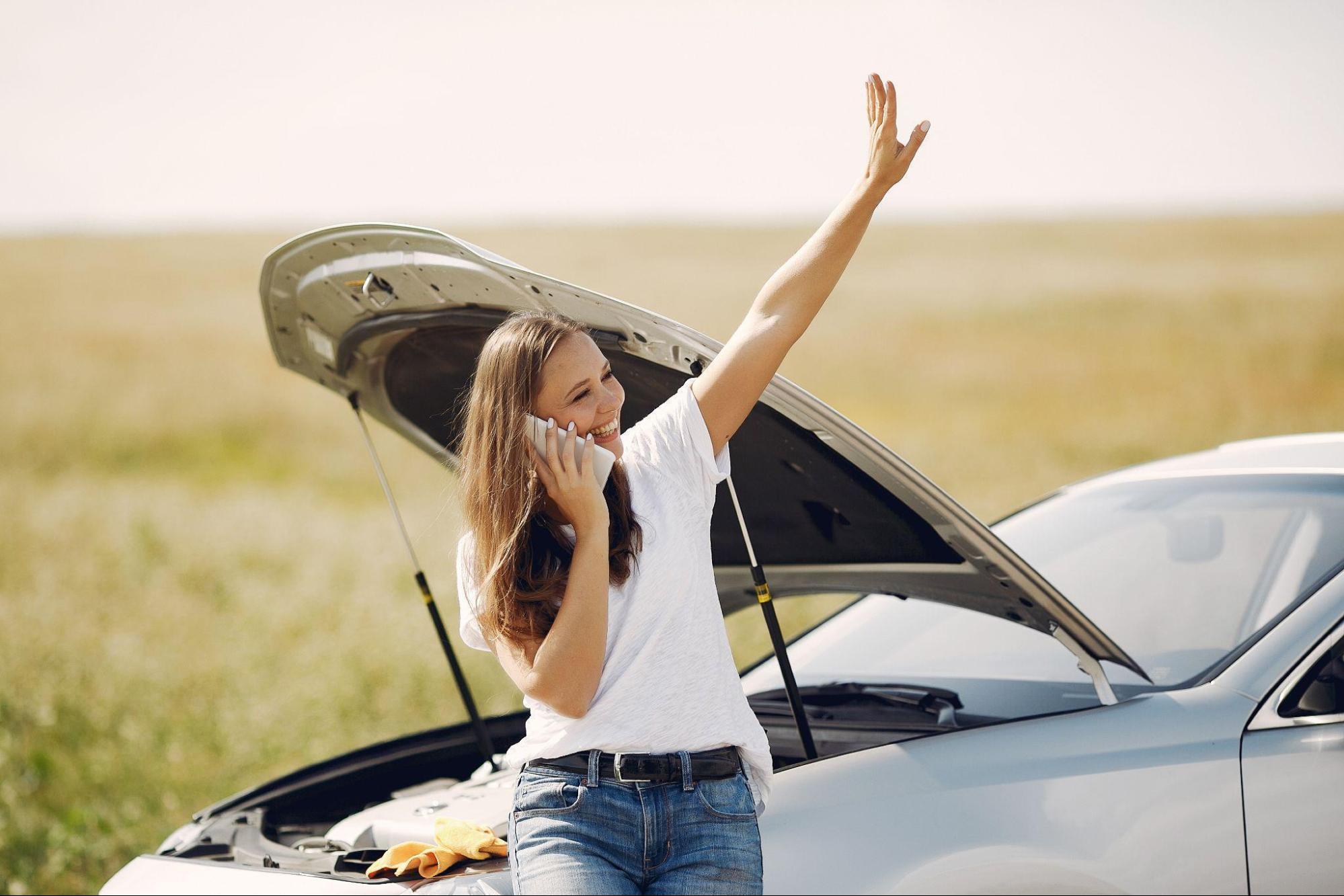 Mulher usando blusa branca básica e calça jeans, parada a beira de uma estrada com o capô do seu carro aberto.