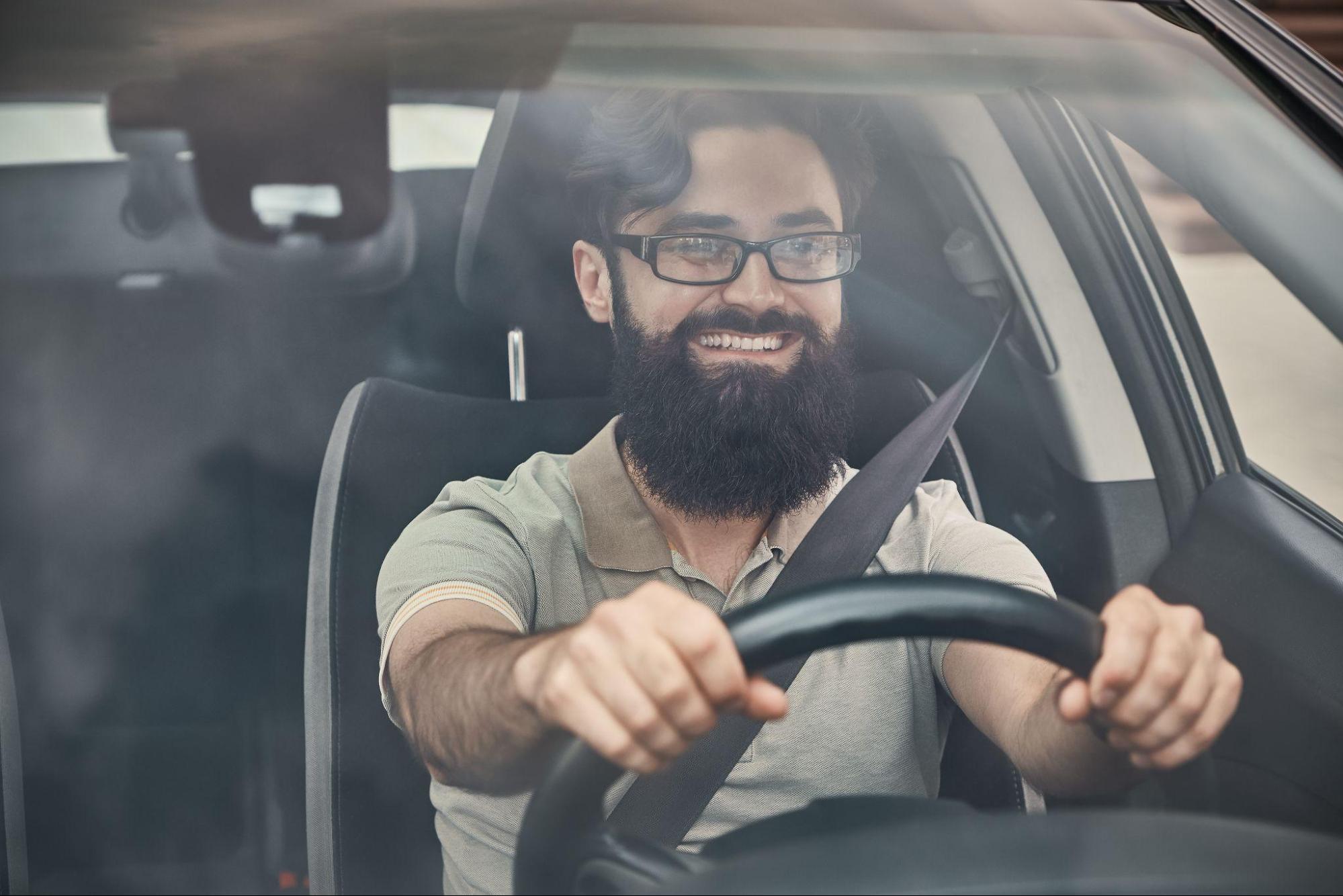 A imagem mostra um rapaz, usando uma camisa cinza e óculos de grau, dirigindo seu carro.