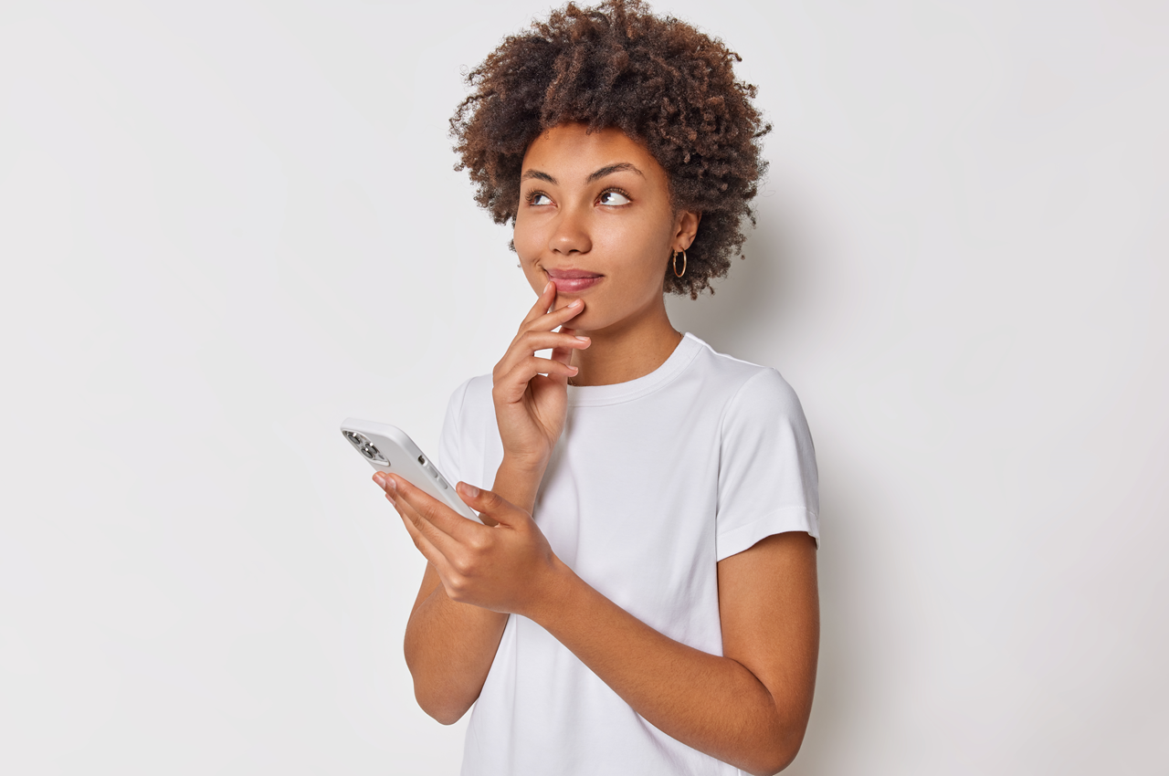 Mulher pensativa com expressão curiosa, segurando um smartphone. Ela veste uma camiseta branca e está em frente a um fundo claro.