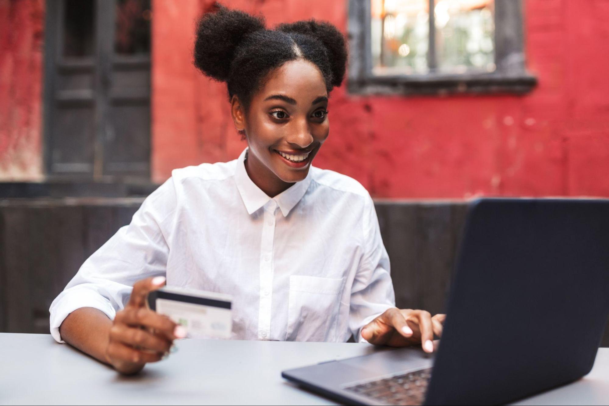 Mulher sorridente em frente a um laptop, segurando um cartão de crédito. Ela veste uma camisa branca e está em um ambiente externo, com uma parede vermelha ao fundo.