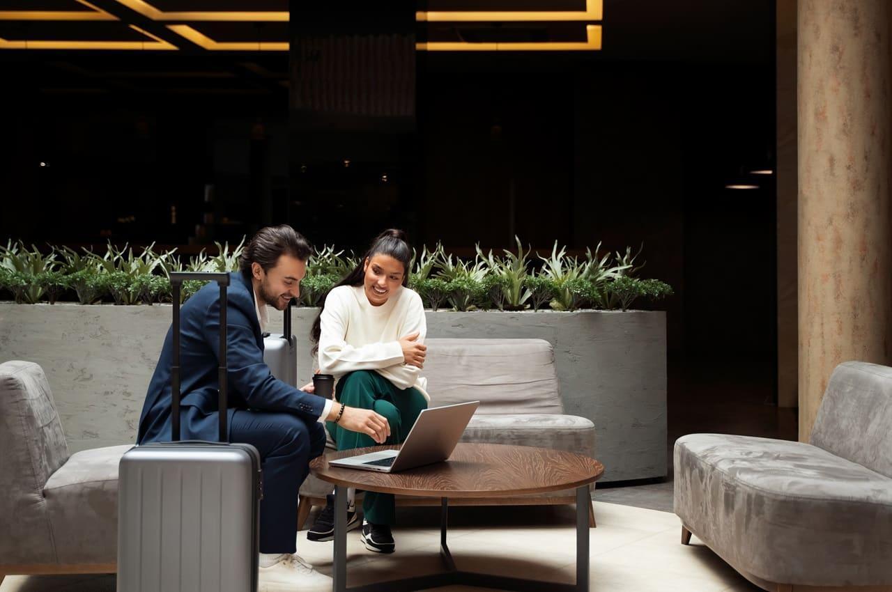 Um homem e uma mulher sentados em um lounge, sorrindo enquanto olham para um laptop. O homem tem uma mala de viagem ao lado, e os dois parecem envolvidos em uma conversa animada. Ao fundo, há plantas decorativas e um ambiente bem iluminado.