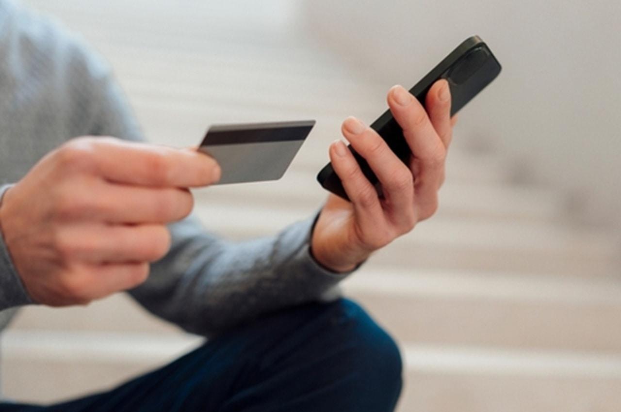 Homem segurando um cartão de crédito com uma mão e um smartphone com a outra. Ele parece estar sentado em escadas. O foco da imagem está nas mãos e nos objetos, com um fundo desfocado.