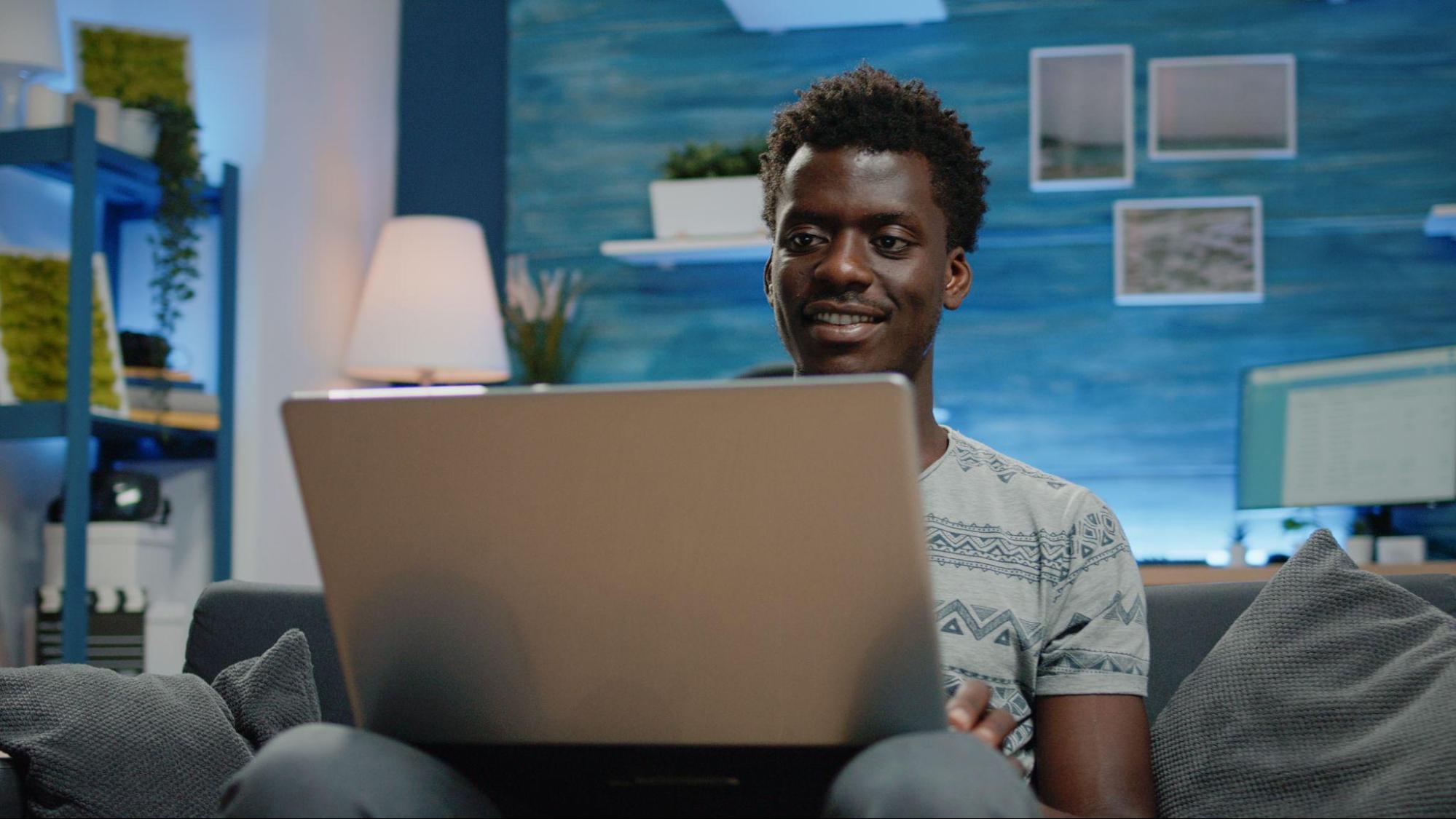 A imagem mostra um rapaz sorridente, usando uma camiseta azul e usando seu laptop.