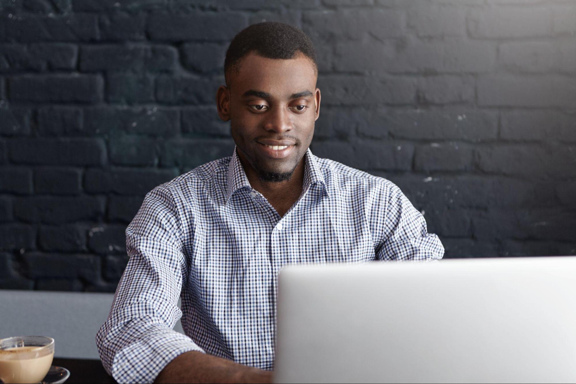 Homem usando camisa social xadrez em um escritório. Ele está sentado e usando seu notebook.