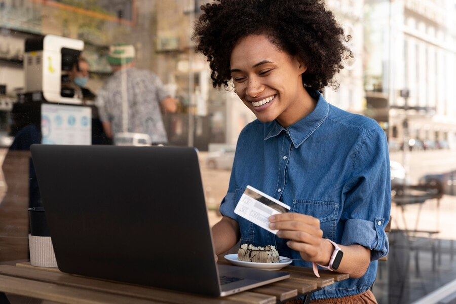 Mulher negra de cabelos crespos sentada em um café, sorrindo, com um notebook à sua frente. Ela usa camisa jeans e segura um cartão de crédito com uma das mãos.