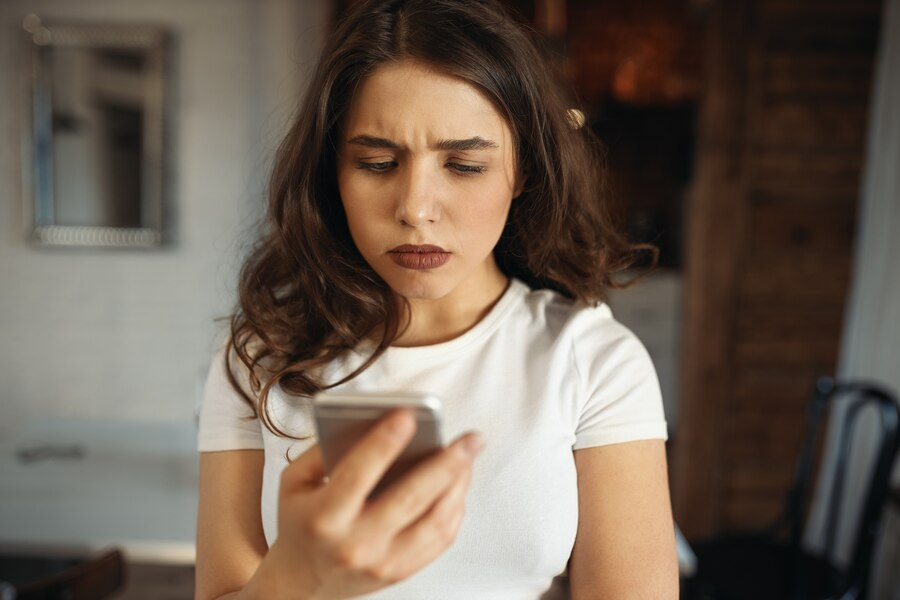 Uma mulher jovem, de pele branca e cabelos castanhos ondulados, segura um celular e o encara com feição preocupada. Ela usa uma blusa branca e batom cor vinho.