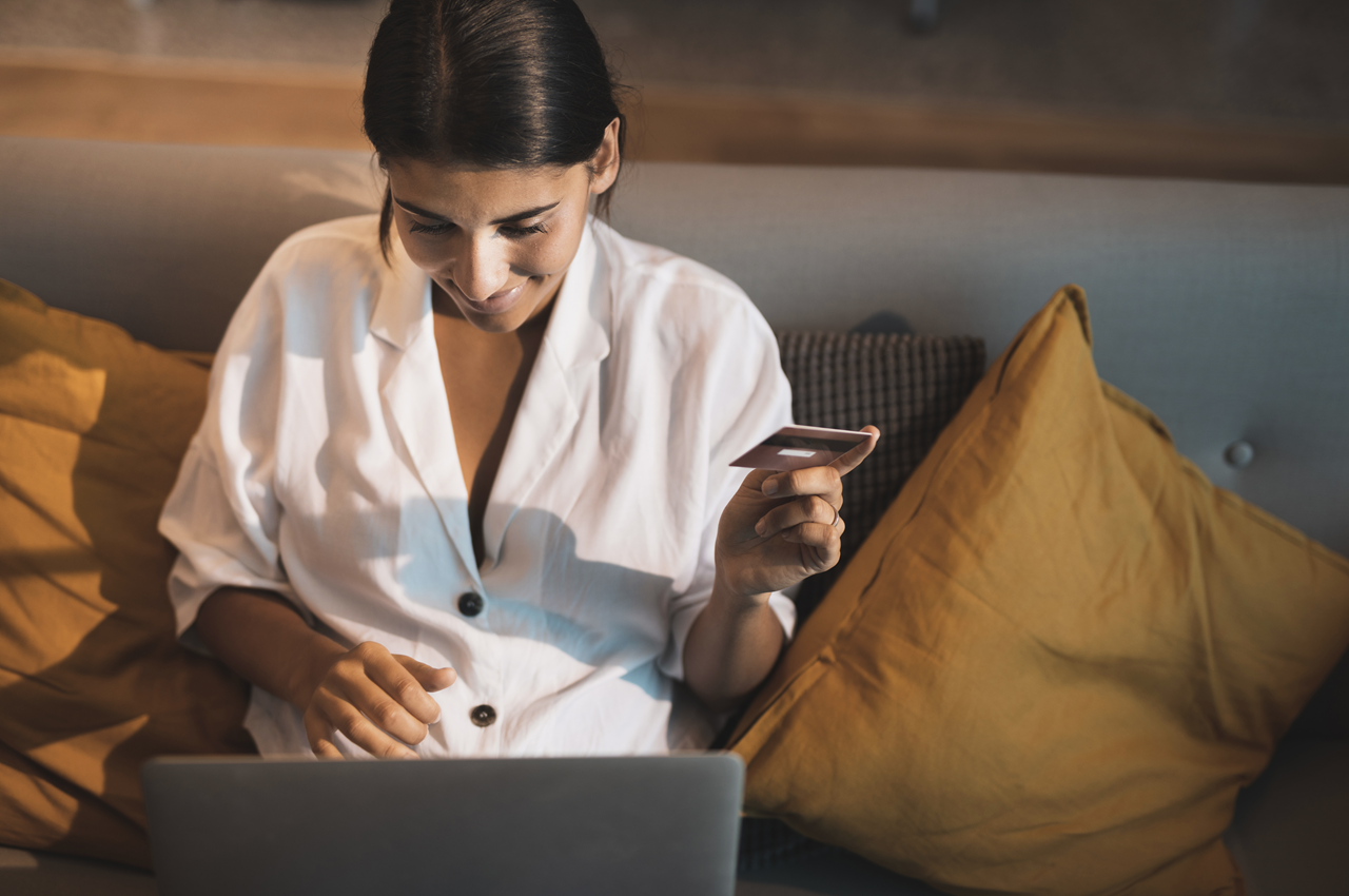 Mulher sorrindo enquanto segura um cartão de crédito em uma mão e usa seu laptop com outra, sentada em um sofá com almofadas amarelas.