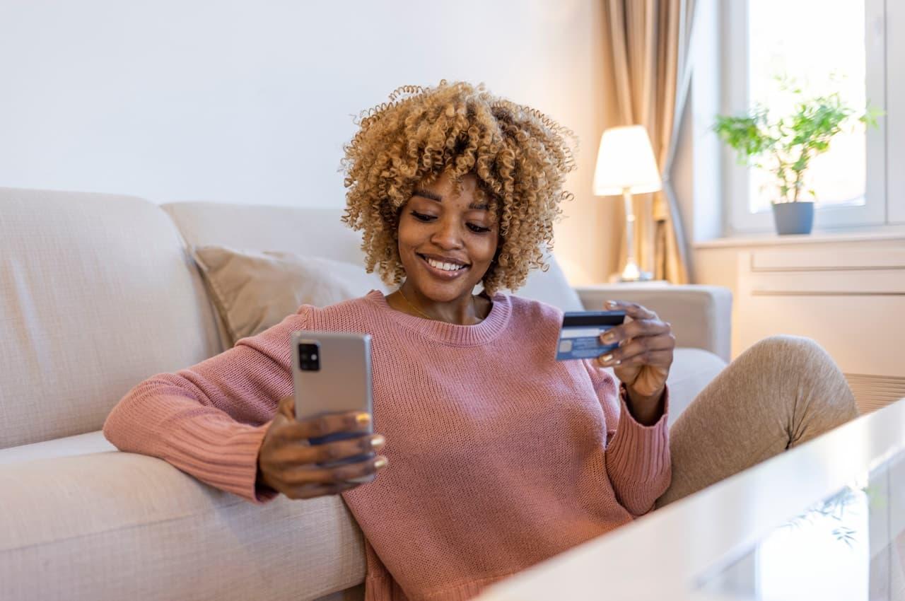 Mulher sentada no chão e encostada em seu sofá. Em uma das mãos ela está usando seu celular e na outra segurando um cartão de crédito.