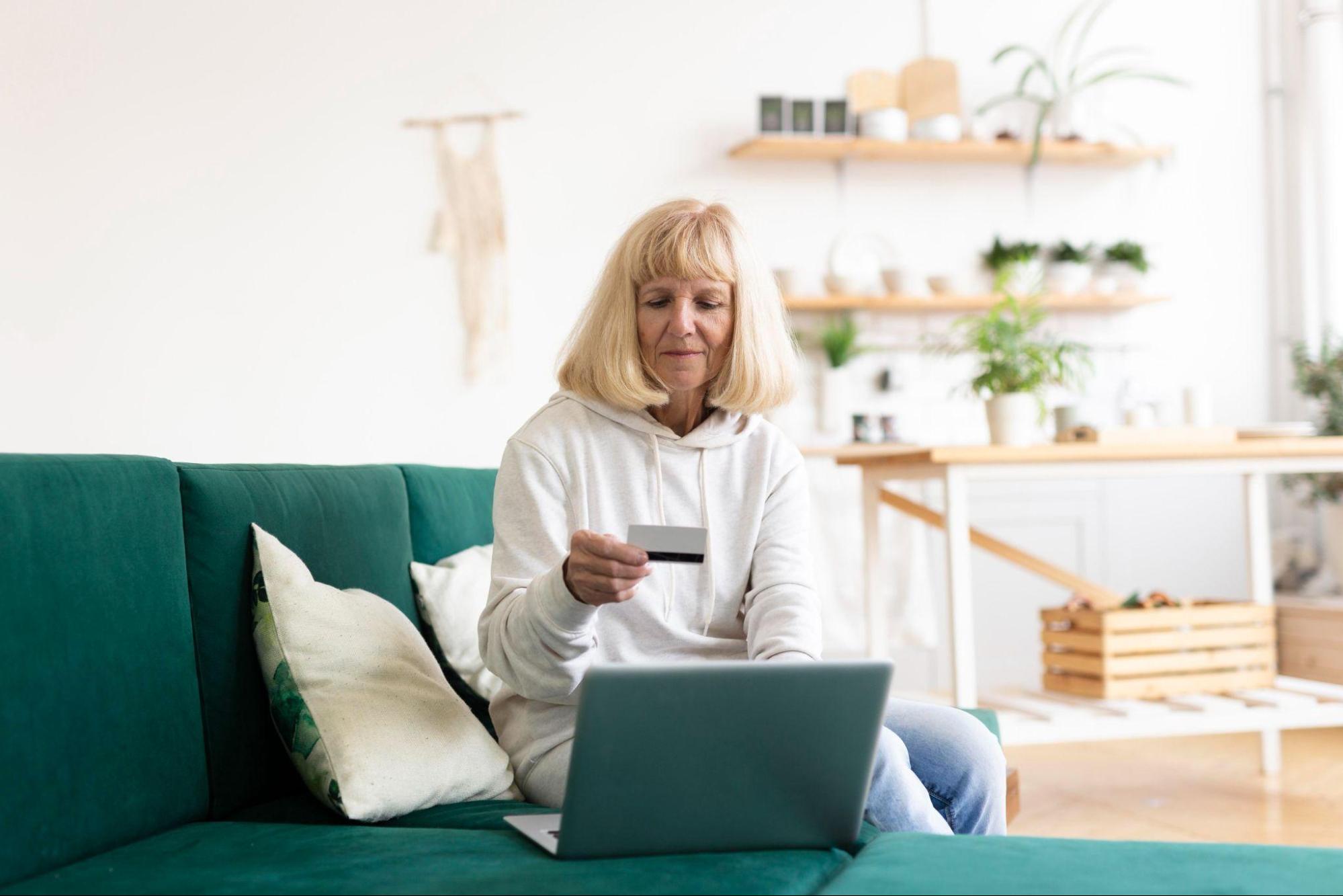 Mulher sentada em um sofá verde, segurando um cartão de crédito em frente a um laptop. Ela está em uma sala de estar com decoração minimalista e plantas ao fundo.