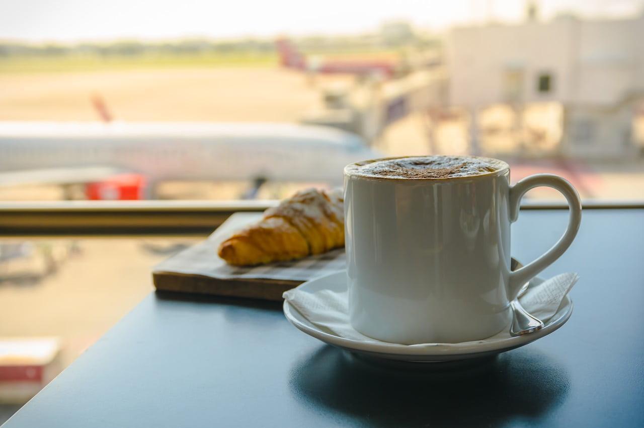 Uma xícara de café em uma mesa, ao lado de um croissant. Ao fundo, é possível ver a pista de um aeroporto e parte de um avião.