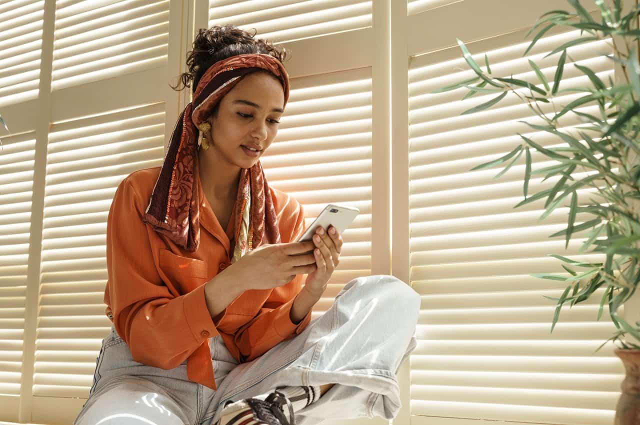 Mulher com cabelo cacheado preso por um lenço, segurando um smartphone. Ela veste uma blusa laranja e está próxima a uma janela com persianas, em um ambiente iluminado.