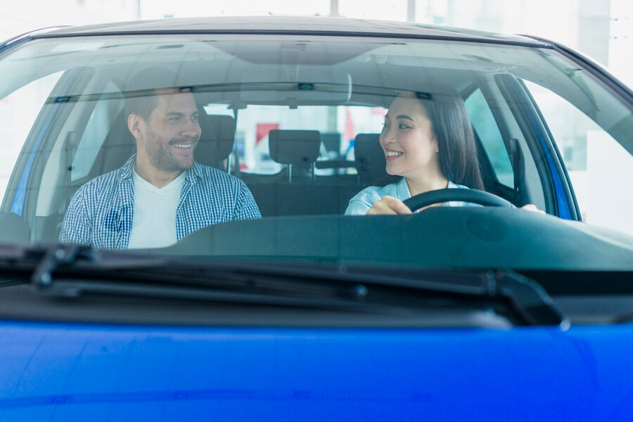 Um homem e uma mulher estão dentro de um carro azul em uma concessionária. O homem está sorrindo enquanto olha para a mulher. A cena é vista pela perspectiva do capô do carro.