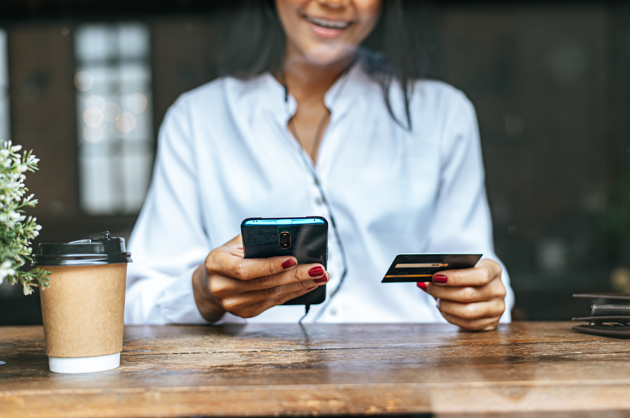 Mulher sorrindo enquanto segura um telefone celular na mão esquerda e um cartão de crédito na mão direita. Ela está sentada à mesa com um copo de café e uma planta ao lado.