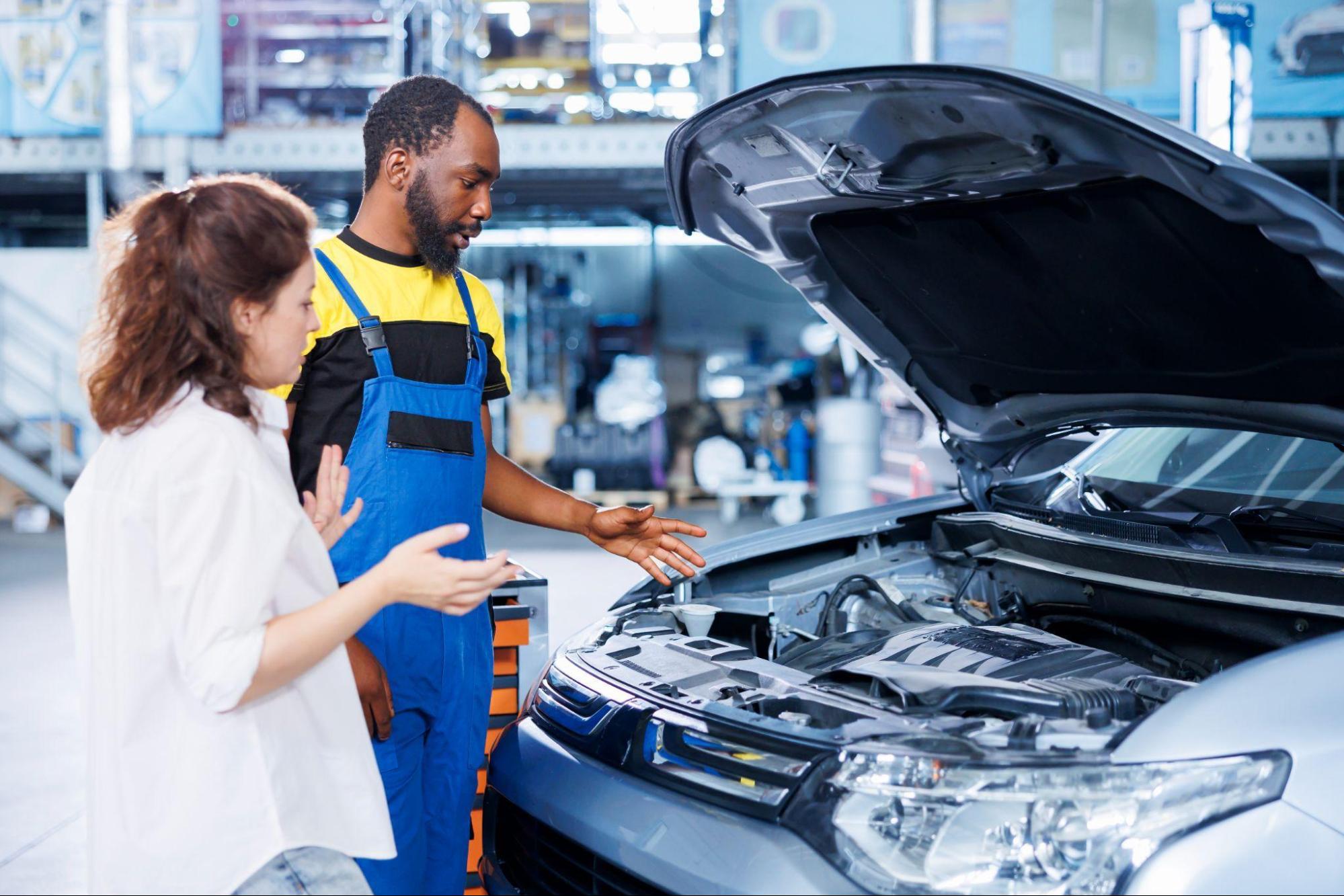 A imagem mostra um rapaz mecânico e uma mulher olhando o motor de um carro.